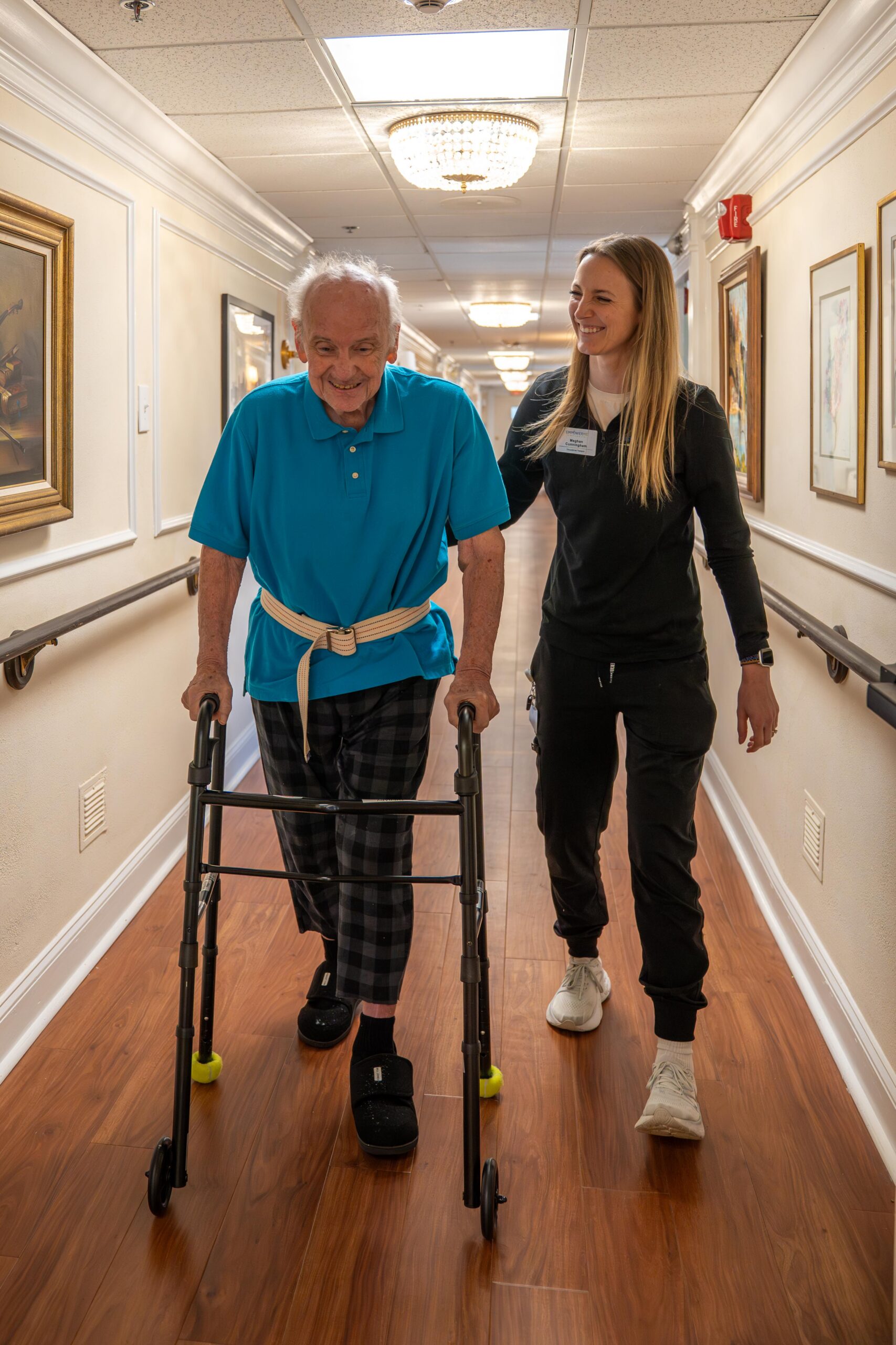 Caregiver helping an elderly memory care resident walk safely down a bright hallway